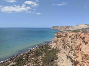 A scenic coastal view featuring rugged cliffs with layers of reddish-brown rock, lush green vegetation near the edges, and the expansive blue ocean under a partly cloudy sky. The horizon stretches, seamlessly blending water and sky in the distance.