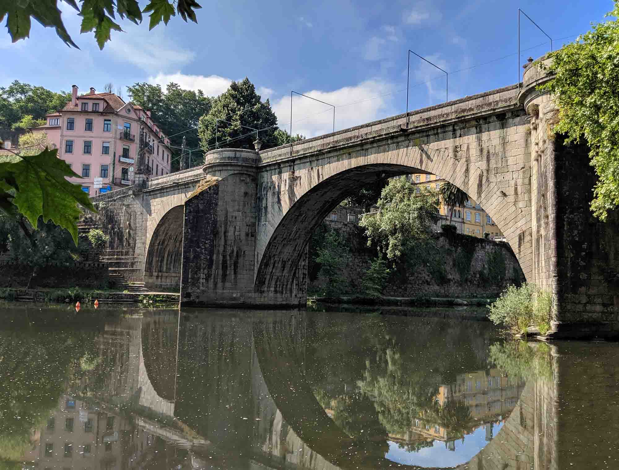 A stone arch bridge spans over a calm river, with its reflection visible in the water. In the background, multi-story buildings reminiscent of Portugal's architecture nestle among green trees under a partially cloudy sky. Branches and leaves frame the scene at the top left corner.