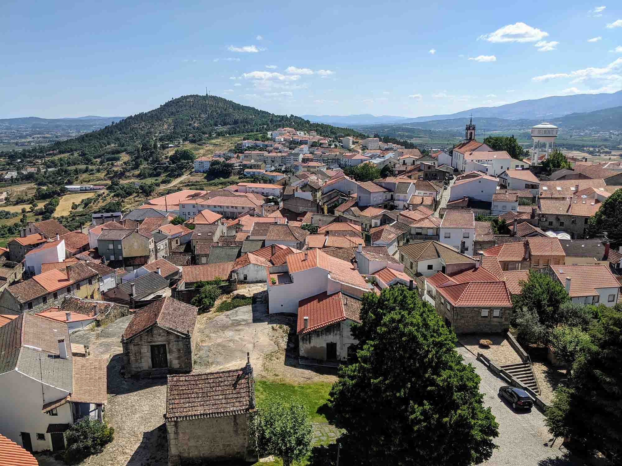 A picturesque view of a small Portuguese town with red-tiled roofs packed closely together. The town is surrounded by rolling hills and lush greenery under a clear blue sky with a few scattered clouds. A prominent hill towers in the background.