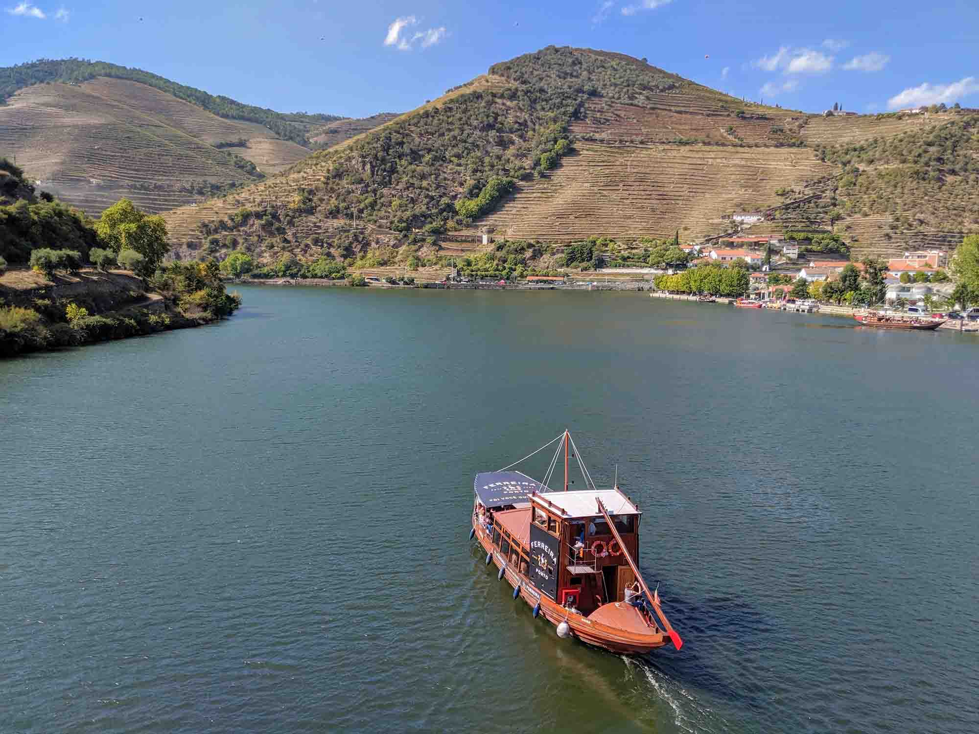 A traditional wooden boat sails on the Douro River in Portugal, surrounded by terraced vineyards and rolling hills. The landscape features lush greenery and quaint buildings, bathed in sunlight under a clear blue sky.
