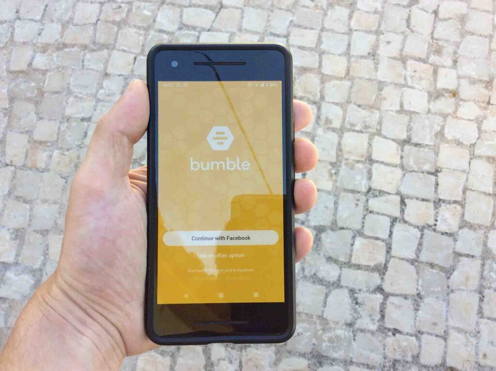 A hand holds a phone with Bumble login options, set against Portugal-style stone pavement suggesting a Portuguese street scene.