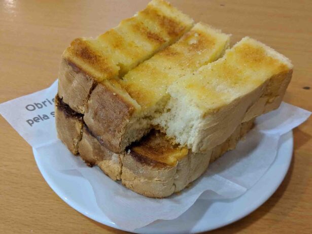 A plate with thickly sliced pieces of buttered toast stacked on top of each other. The toast appears golden brown on top, with a piece slightly pulled away revealing the soft, fluffy interior. The plate rests on a wooden table with a napkin underneath.