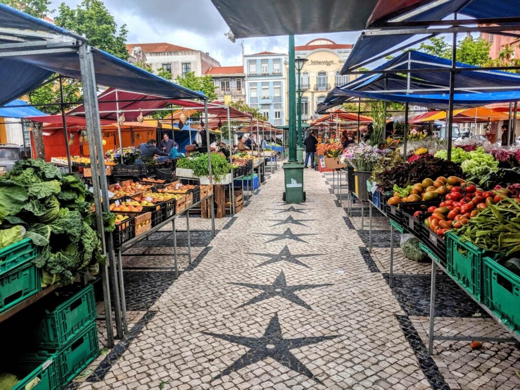 Outdoor market scene with colorful stalls on both sides displaying various fruits, vegetables, and leafy greens. The ground is paved with decorative tiles featuring star patterns, reminiscent of Portugal. People are seen browsing the stalls under multi-colored awnings; background shows buildings.