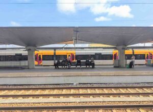 A train station platform with several people sitting on benches under a canopy, waiting beside the yellow and white train. Train tracks run parallel in the foreground, and the sky is partly cloudy.