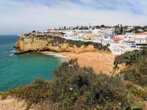 A picturesque coastal scene shows a sandy beach surrounded by rugged cliffs and clear blue waters. White-washed buildings with red and yellow roofs overlook the beach from the cliffs above. People are sunbathing and enjoying the beach. Small plants are in the foreground.