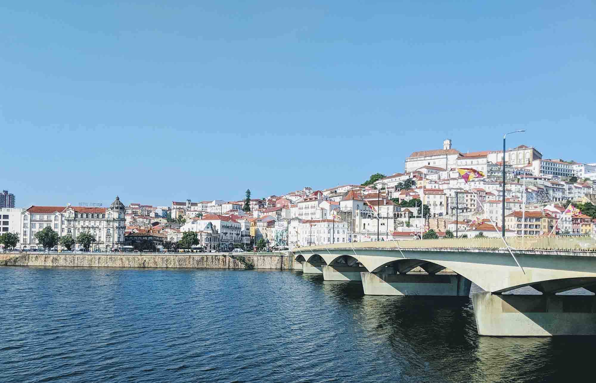 A picturesque cityscape in Portugal with numerous white and orange-roofed buildings on a hill, partially framed by a bridge crossing a calm river under a clear blue sky.