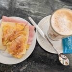 A plate with a sandwich made of ham, cheese, and a croissant sits next to a glass of frothy coffee on a saucer with a spoon, and two packets of sugar on a dark marbled table.