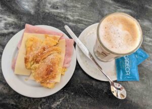 A plate with a sandwich made of ham, cheese, and a croissant sits next to a glass of frothy coffee on a saucer with a spoon, and two packets of sugar on a dark marbled table.