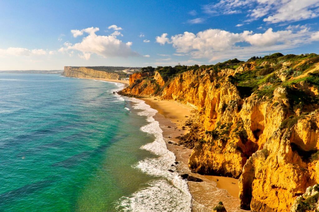 Golden cliffs border a sandy Portuguese beach with turquoise waves, lush cliffside plants, and a partly cloudy sky overhead.
