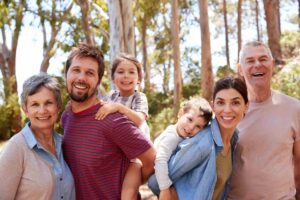 A multi-generational family, composed of two grandparents, two parents, and two young children, poses outdoors in a forested area. They are all smiling and appear happy. The children are being carried by the adults. Trees and sunlight are visible in the background.