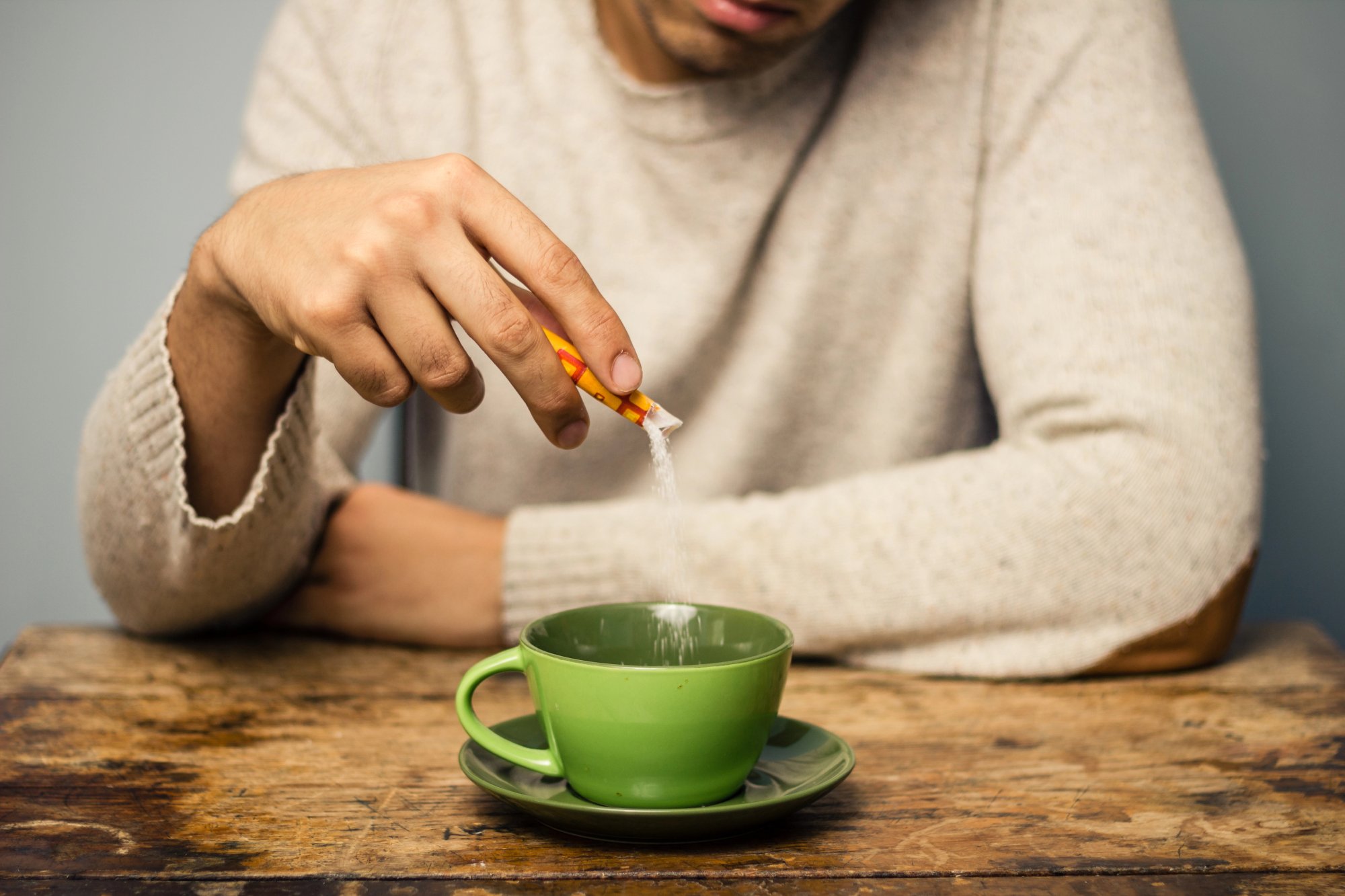 A person in a beige sweater pours sugar from a packet into a green cup and saucer on a wooden table. The person's face is partially out of frame.