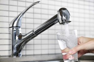 A hand is holding a clear glass under a chrome kitchen faucet, filling it with water. The background features white tile walls.