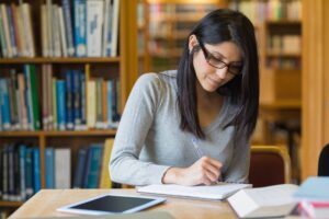 A woman with glasses, wearing a grey sweater, is seated at a wooden table in a library. She is writing in a notebook, with several open books and a tablet beside her. Bookshelves filled with books are visible in the background.