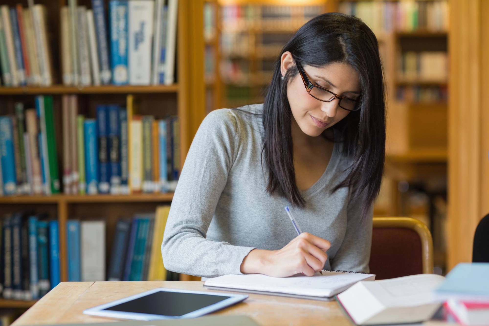 A woman with glasses, wearing a grey sweater, is seated at a wooden table in a library. She is writing in a notebook, with several open books and a tablet beside her. Bookshelves filled with books are visible in the background.