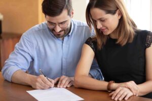 A man and a woman sit at a table, reviewing and signing a document together. The man, wearing glasses and a light blue shirt, holds a pen and signs the paper, while the woman in a black dress with lace sleeves looks on attentively.