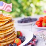 A stack of pancakes topped with melting butter, syrup, and a small American flag on a plate. Fresh strawberries and blueberries garnish the plate. Additional bowls of blueberries and strawberries are in the background. The scene is set outdoors.