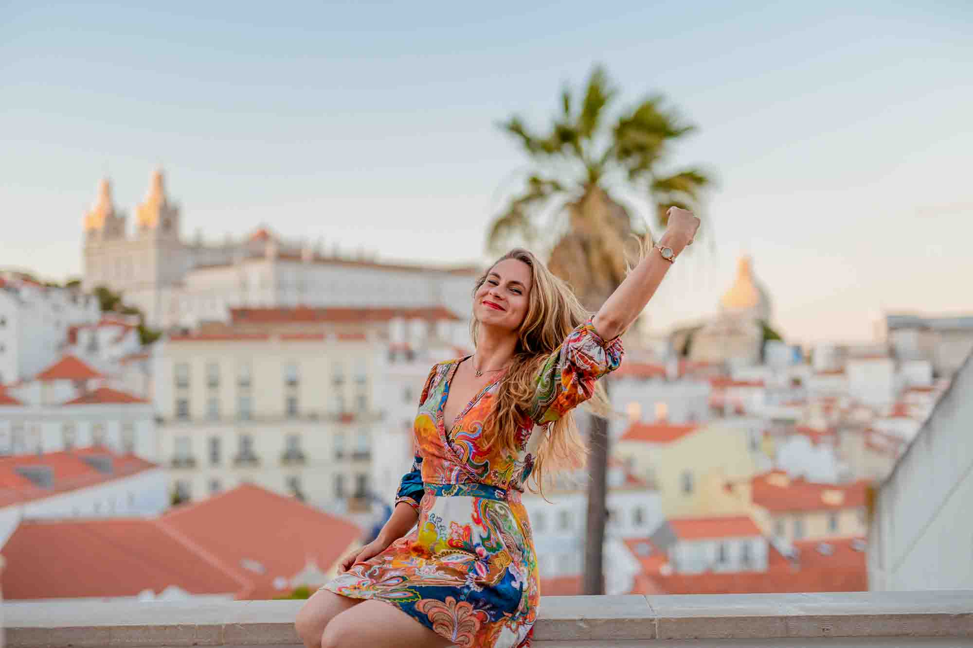 A woman wearing a colorful dress sits on a ledge with her right arm raised and smiling. She is outdoors, with a scenic backdrop of buildings with red rooftops and palm trees under a clear sky.