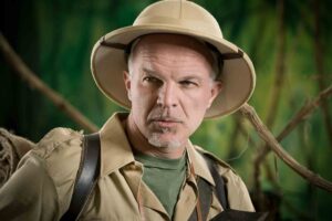 A person wearing a safari hat and khaki clothing stands in front of a jungle-like backdrop, gazing intently at the camera. They have a serious expression, with a determined look on their face. The background is lush and green, giving a sense of exploration.
