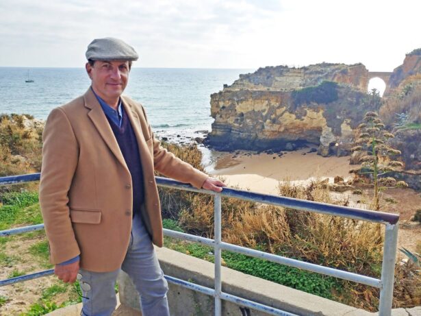 A man in a beige coat and gray flat cap stands by a metal railing overlooking a scenic coastal landscape with cliffs, a sandy beach, and a sea arch. There is greenery around him and a boat can be seen in the distance on the water.