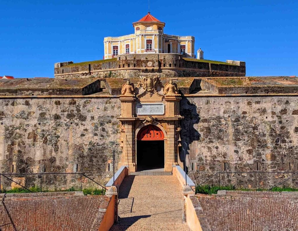 The image showcases the citadel of Elvas, Portugal, with a large stone wall and an ornate archway entrance. The fort is perched on a hill, with a well-preserved, historic building featuring a red roof at its apex under a clear blue sky.