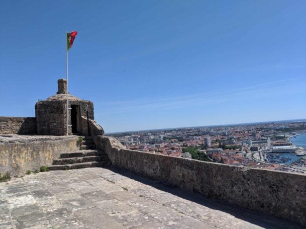 A stone structure with a Portuguese flag atop stands on a high vantage point overlooking a sprawling Setubal cityscape and harbor in Portugal. The sky is clear and blue, enhancing the visibility of the town's red-roofed buildings, waterfront, and distant horizon.