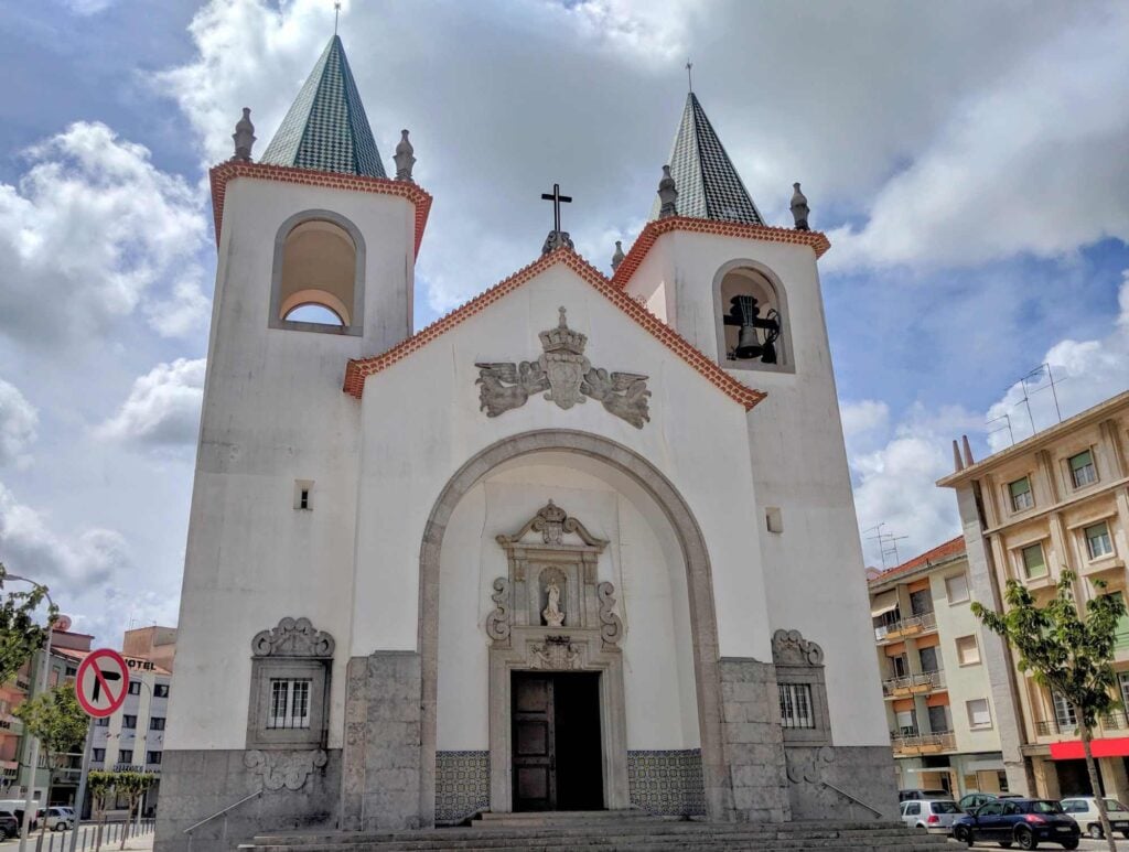 A church with twin bell towers, green-tiled roofs, and a central arched entrance featuring a sculpture above the wooden doors. The building is flanked by streets with parked cars and neighboring buildings. The sky is partly cloudy, reminiscent of architectural styles found in Portugal.