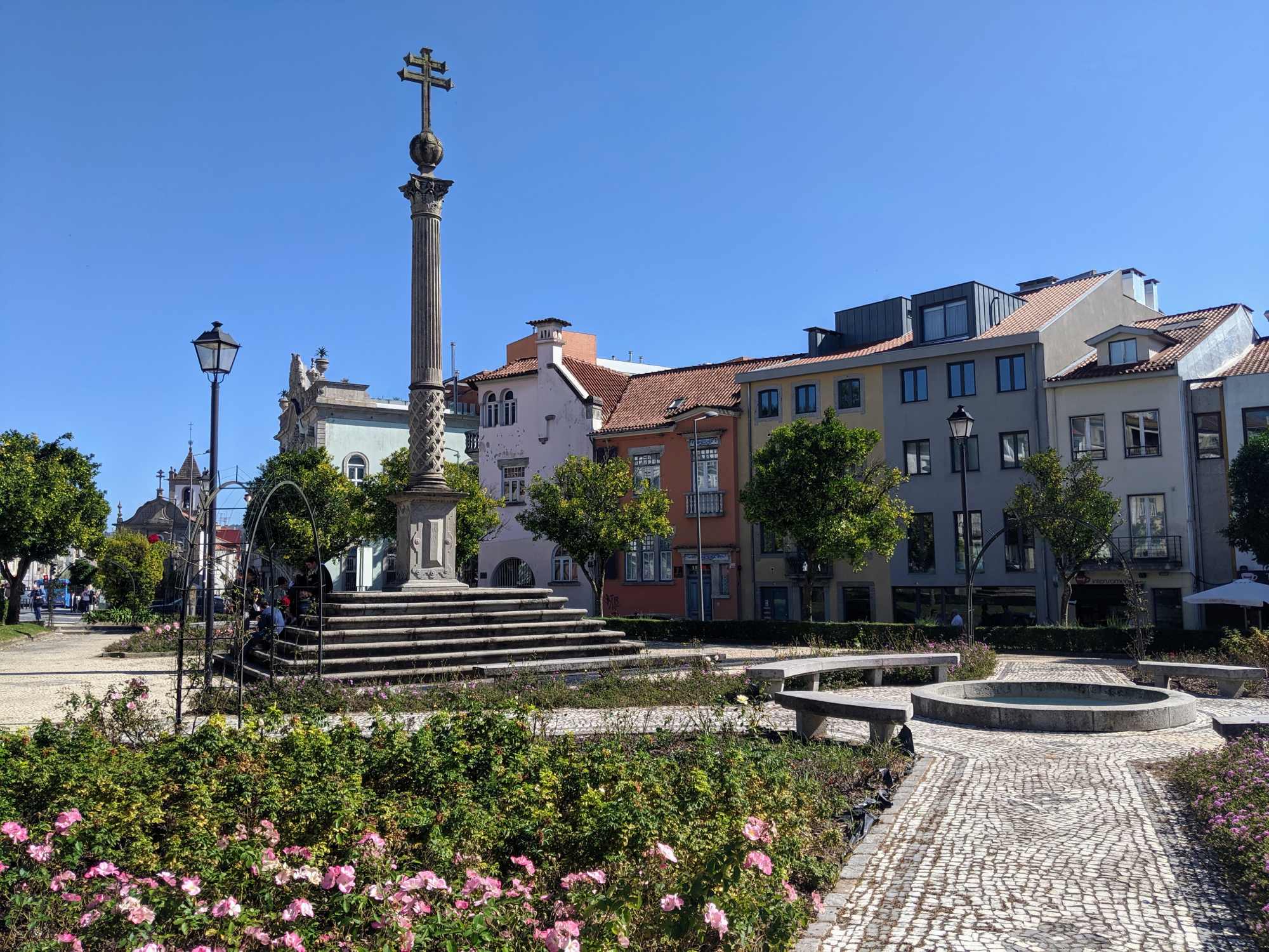 A sunny town square in Braga, Portugal features a stone monument with a cross on top, surrounded by a garden of blooming pink flowers. Colorful multi-story buildings with traditional architecture line the background, and a few people sit near the monument.