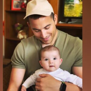 Joel wearing a beige cap and green t-shirt is holding a baby dressed in white. The man is looking down and smiling at the baby, who is staring at the camera with a curious expression. They are indoors, with a shelf and a television in the background.