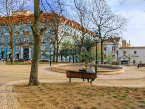 Two people sit on a park bench facing a circular fountain in a city square. The park is surrounded by bare trees and colorful buildings with a blue and white facade, reminiscent of Portugal's charming architecture. It's a clear day with a few people walking in the background.