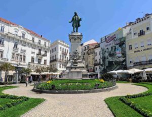 A historical town square with a central statue atop a tall pedestal surrounded by a circular garden with yellow flowers. The background features colorful multi-story buildings, outdoor café seating, and people walking under a bright blue sky.