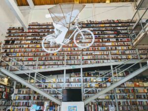 A large library in Portugal with shelves filled with books, a metal staircase leading to an upper level, and a white sculpture of a person riding a bicycle suspended in mid-air from the ceiling.