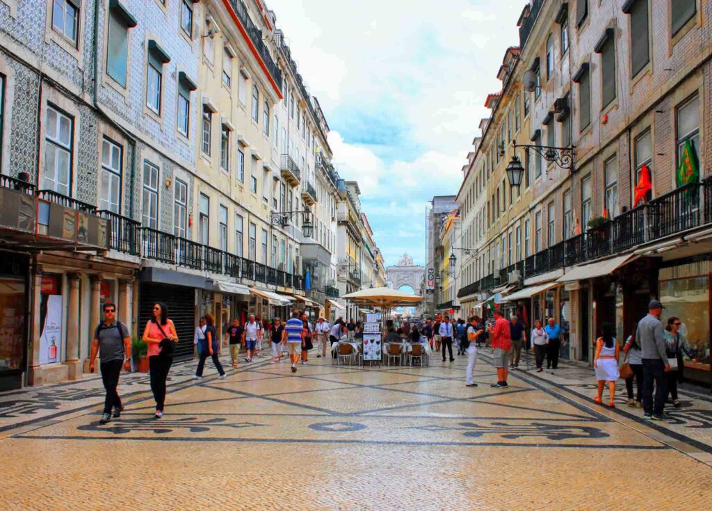 A bustling European street lined with historic buildings featuring ornate balconies and a mix of shops and cafés. Pedestrians stroll along the cobblestone pavement with intricate black and white patterns, reminiscent of Portugal’s iconic designs. In the distance, a café with outdoor seating is visible.