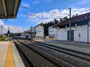 A small train station with two rail tracks and platforms. The station building is white with a red-tiled roof. There are electric lines above the tracks and a signal light. The sky is mostly clear with a few clouds, and trees are visible in the background.