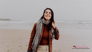 A woman in a brown jacket, gray scarf, and beige pants stands on a foggy beach smiling at the camera. The background features a calm, overcast ocean. In the bottom right corner, there's a small logo with text that reads "Learn European Portuguese Online.
