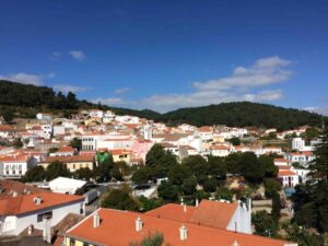 A picturesque town in Portugal with a cluster of white and pastel-colored houses with orange-tiled roofs nestled among lush green hills under a clear blue sky. The vibrant, serene landscape features a mix of residential buildings and greenery.