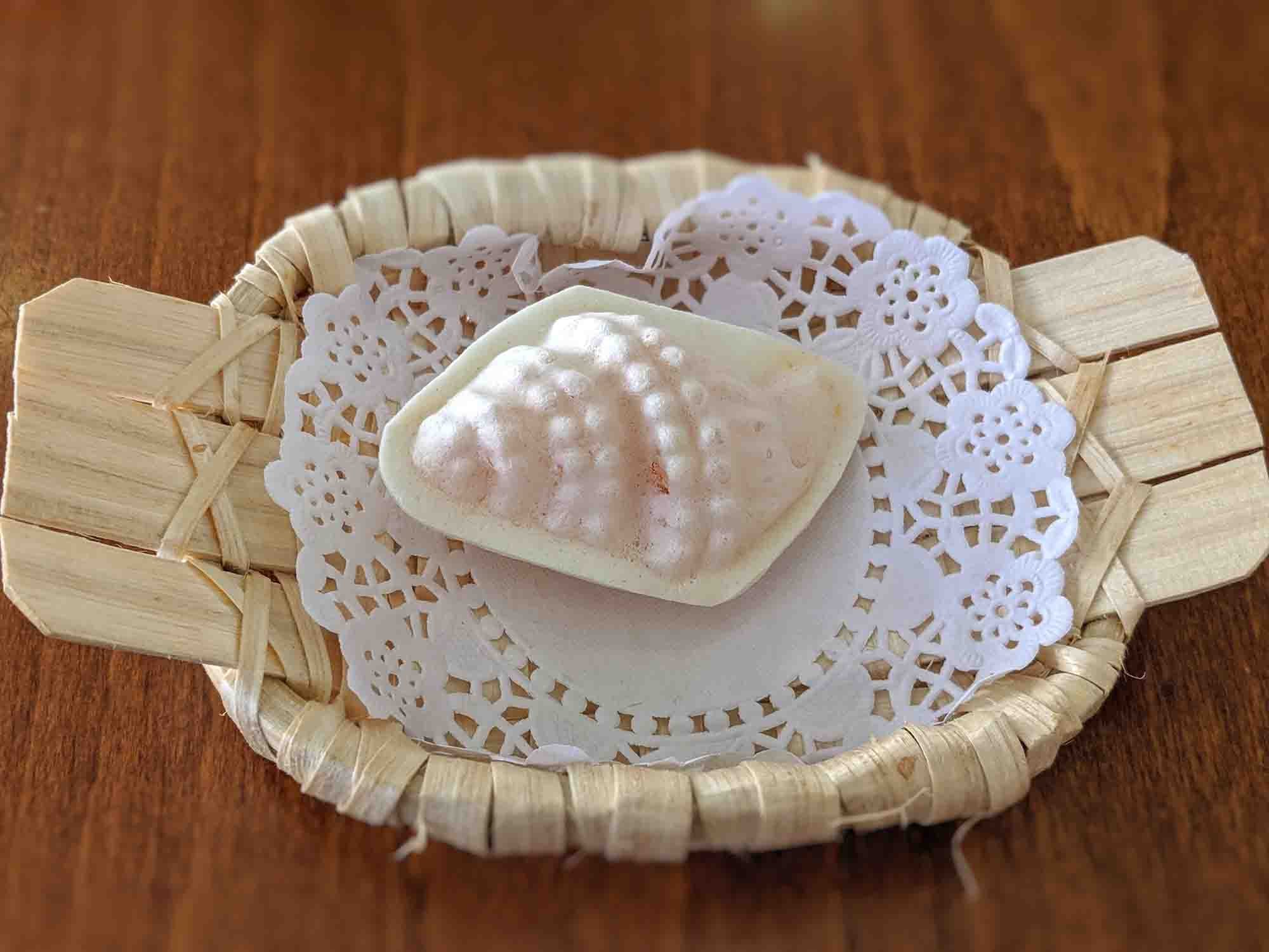 A piece of traditional Indian sweet called khaja, characterized by its pale color and irregular bubbled texture, is presented on a white doily in a small woven basket with a rustic wooden background, reminiscent of charming Portuguese cafés. A piece of traditional Indian sweet called khaja, characterized by its pale color and irregular bubbled texture, is presented on a white doily in a small woven basket with a rustic wooden background, reminiscent of charming Portuguese cafés.