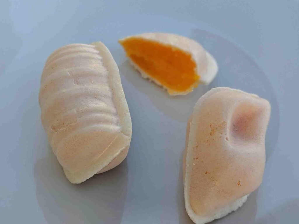 Three pieces of traditional Portuguese pastry are displayed on a white plate. One piece is oval with ridged lines, another is smooth with a slight indent, and the third is cut open, revealing a bright orange filling. The pastries have a soft, white, and fluffy appearance.