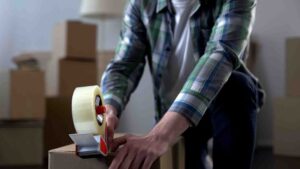 A person wearing a green plaid shirt is crouched down, sealing a cardboard box with clear packing tape using a tape dispenser. Several other cardboard boxes are visible in the blurred background, indicating a moving or packing activity.