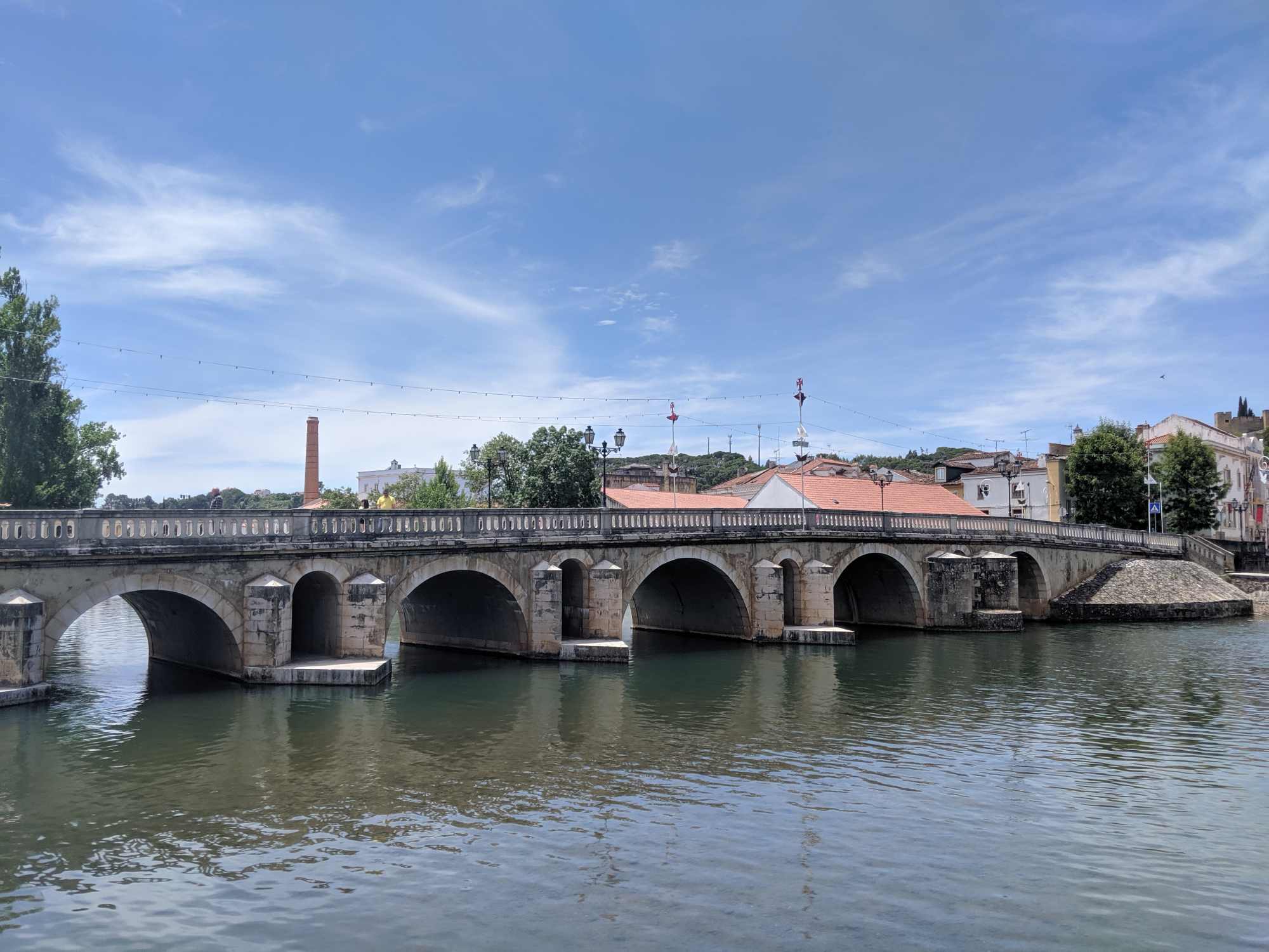 A stone bridge with six arches spans a calm river in Portugal. The bridge connects to a town with buildings featuring red-tiled roofs. Trees and a chimney are visible in the background under a partly cloudy blue sky.