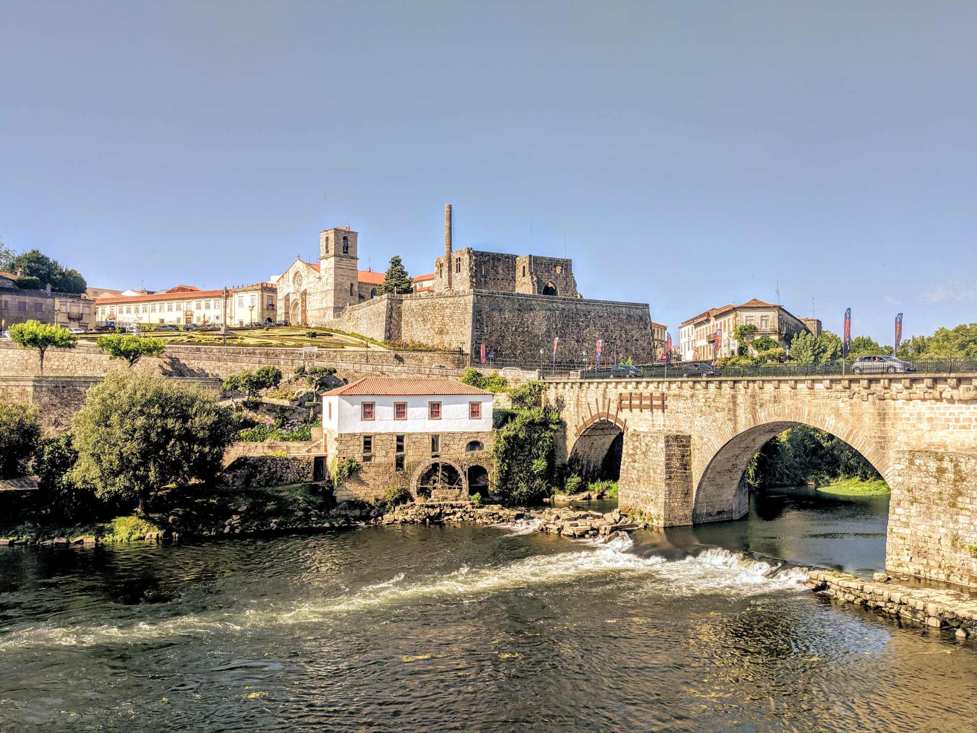 A historic stone bridge spans a river with gentle rapids. On the far side of the river, an ancient fortified complex with a bell tower and large stone buildings stand atop a hill. A small white building is situated near the river’s edge beneath the hill, evoking the timeless charm of Portugal.