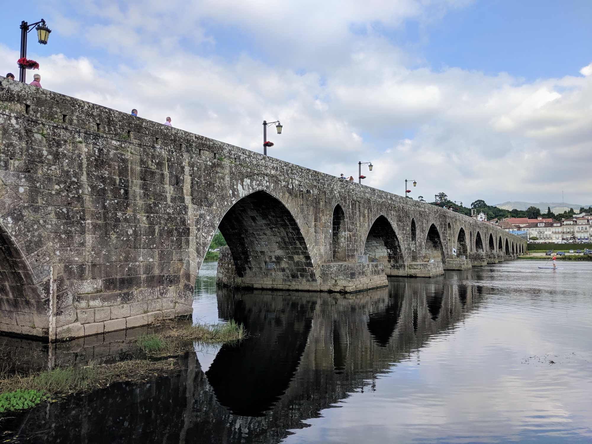 A historic stone bridge with multiple arches spans over a calm river, reflecting its structure in the water. Street lamps line the bridge, and a cloudy blue sky adds to the serene atmosphere. In picturesque Portugal, buildings and greenery complete this charming scene.