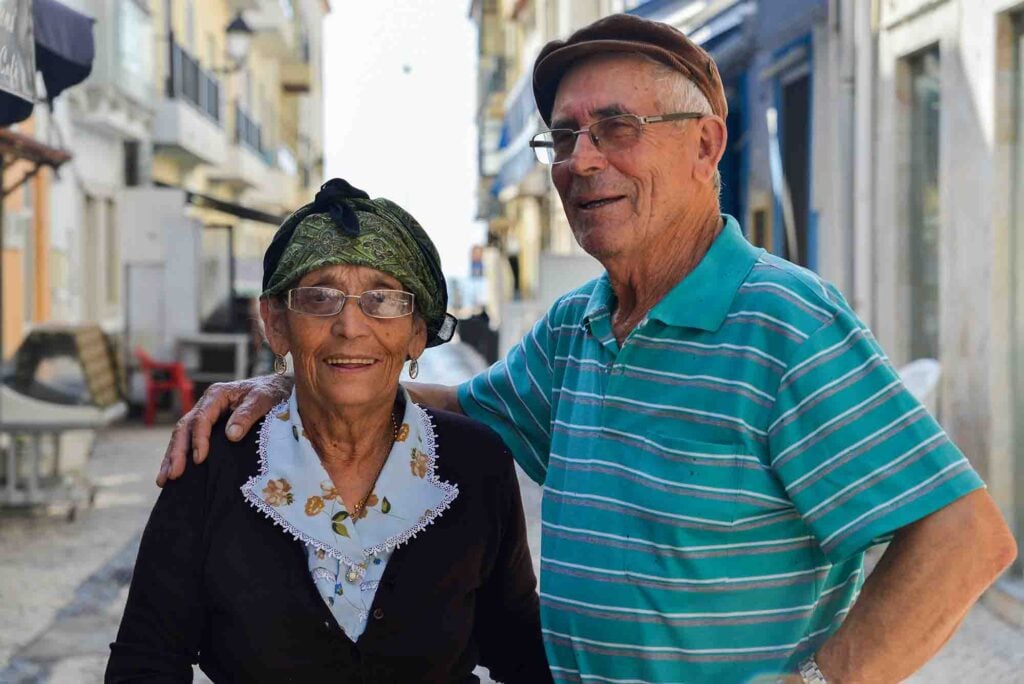 Smiling elderly couple on a sunny street in Portugal; woman with glasses, green scarf, man with brown cap and green striped shirt.