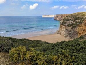 A scenic coastal view featuring a secluded beach surrounded by rocky cliffs and greenery. The calm ocean waves gently lap against the sandy shore under a bright blue sky with a few scattered clouds. Cliffside vegetation adds a touch of green to the landscape.