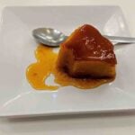 A piece of caramel flan with a glossy top sits on a white square plate. Caramel sauce is spread around the flan, reminiscent of traditional Portuguese desserts. A spoon rests on the plate next to the indulgent treat.