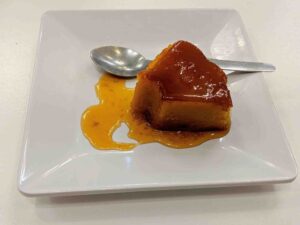 A piece of caramel flan with a glossy top sits on a white square plate. Caramel sauce is spread around the flan, reminiscent of traditional Portuguese desserts. A spoon rests on the plate next to the indulgent treat.