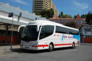 A white and red Rede Expressos bus is parked at a bus station on a sunny day in Portugal. The bus station has a shelter and a few people sitting on benches. In the background, there are trees, residential buildings, and a tall apartment complex.