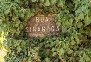 A weathered stone sign partially covered by lush green ivy reads "RUA SINAGOGA." The sign appears to be old, with some letters faded, suggesting it marks a location of historical or cultural significance.