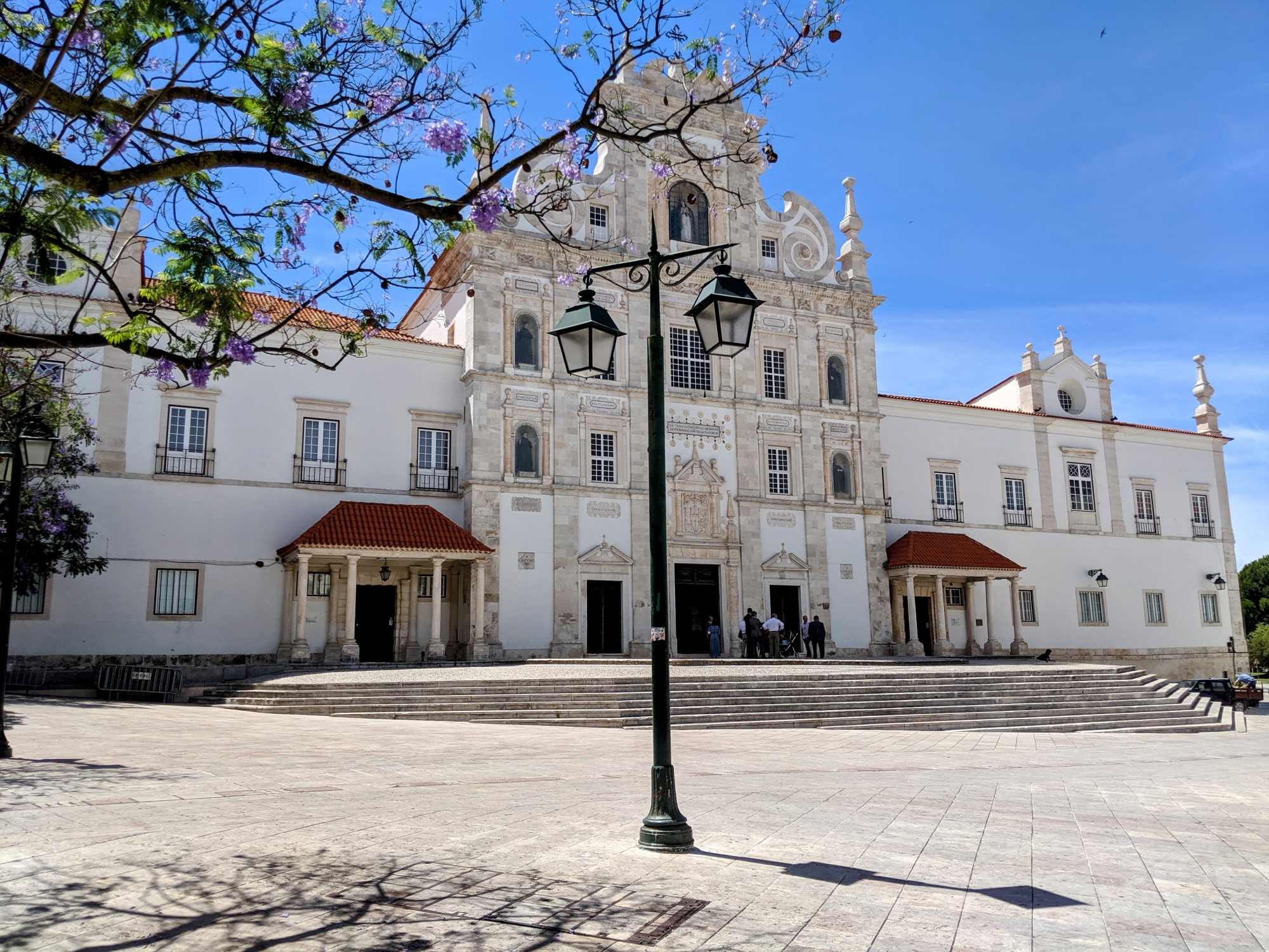 Baroque Portuguese church with white façade and twin towers, purple jacaranda tree, and old streetlamp on a sunny stone plaza.