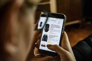 A person is holding a Samsung smartphone, browsing an online shopping app displaying various black jackets. The person appears to be sitting indoors, looking at product images and details on the screen, with a wooden piece of furniture in the background.
