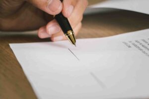A close-up of a hand holding a black and gold pen, signing a document on a wooden surface. The document has text and a signature line visible.